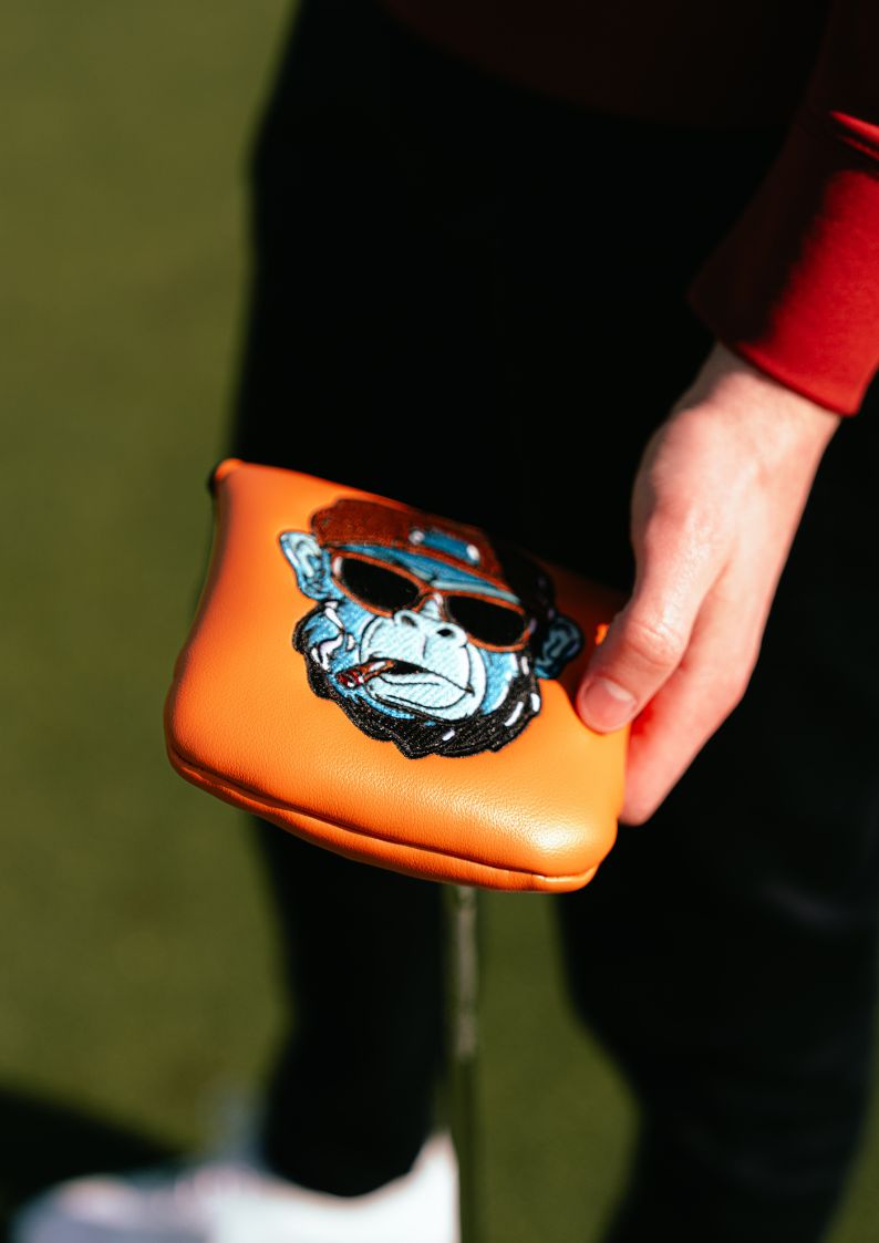 Hand holding an orange golf club head cover with a cartoon character design on a blurred green background.
