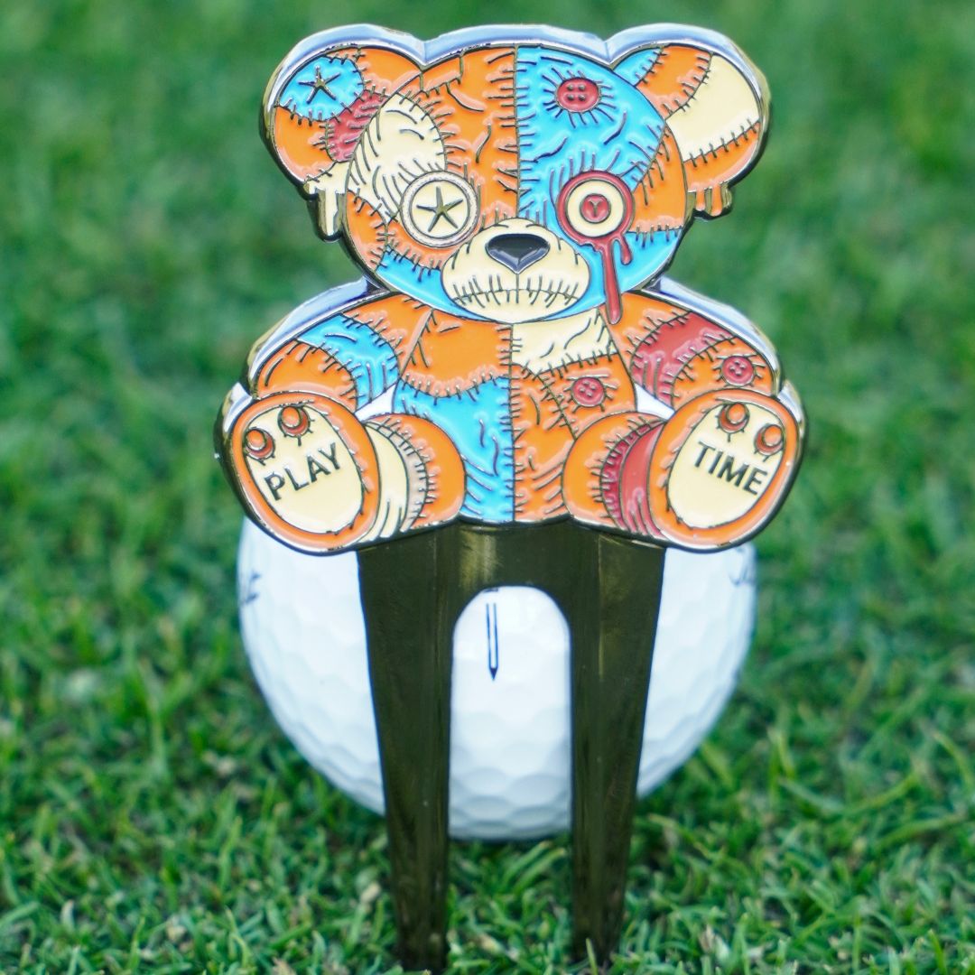 Colorful voodoo bear-shaped golf divot tool in front of a golf ball with grass background