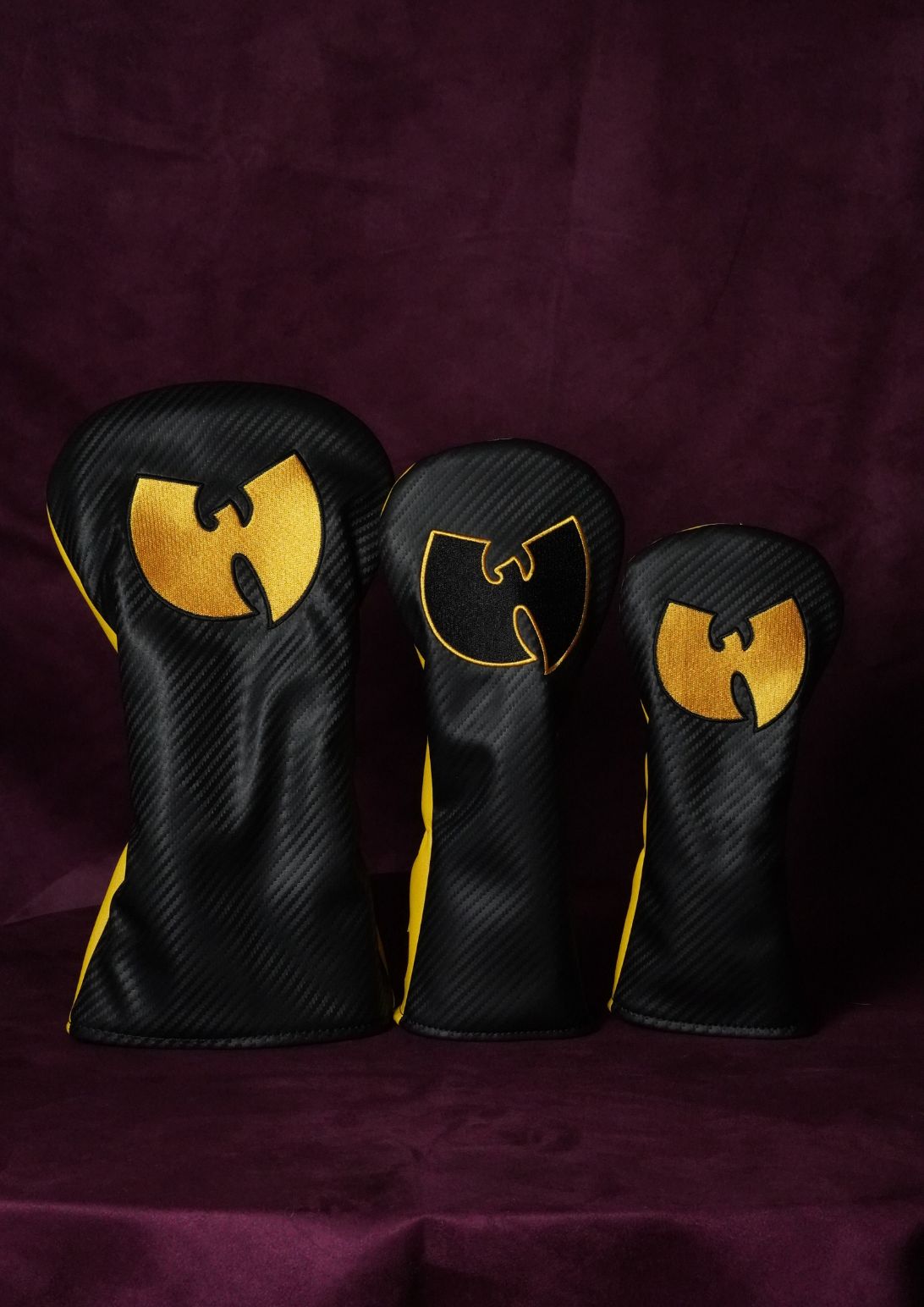 Three black carbon-fiber-textured golf club headcover sleeves with gold Wu-Tang Clan logo designs, arranged in descending size order on a dark purple fabric background.