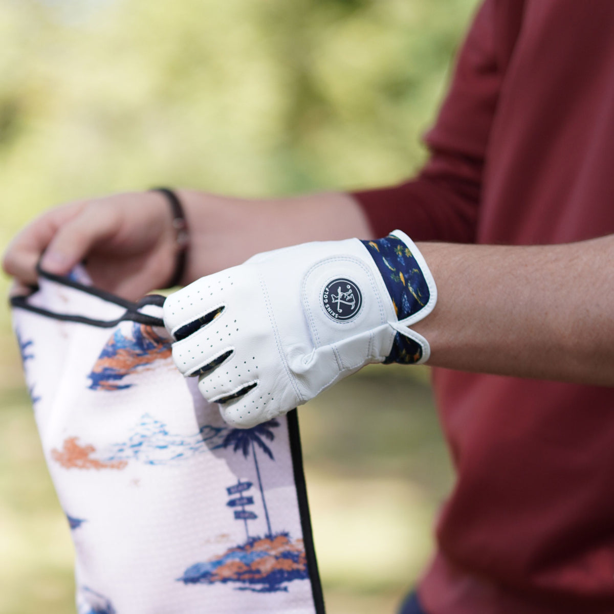 A person outdoors wearing a white golf glove with a circular logo patch, holding a golf towel with a tropical beach print.