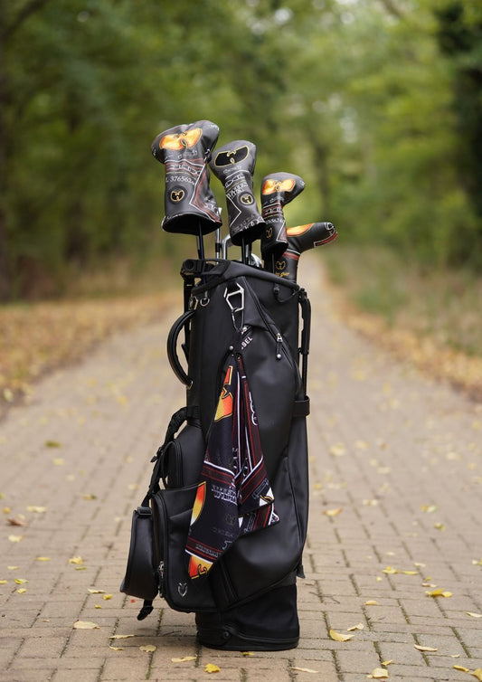 A black golf bag standing on a paved path outdoors, holding several golf clubs with decorative black, orange, and gold headcovers. Trees and fallen leaves are visible in the blurred background.