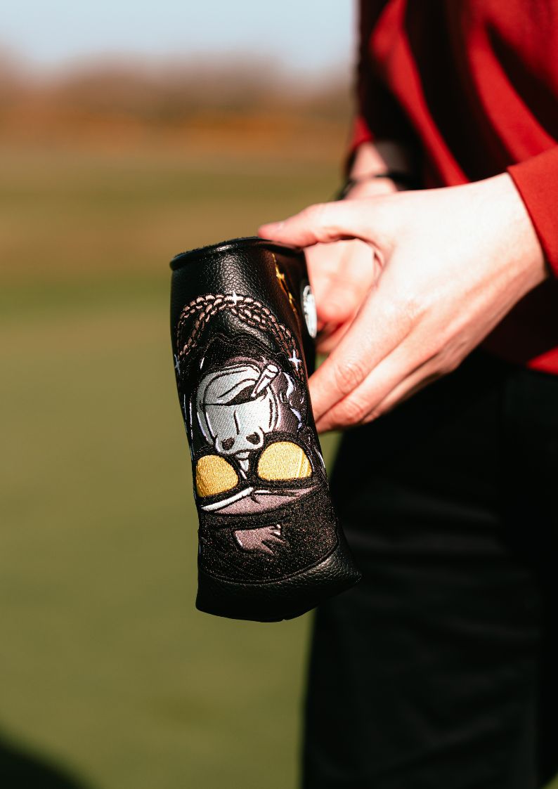 Person holding a black golf headcover with a gorilla design on a blurred outdoor background