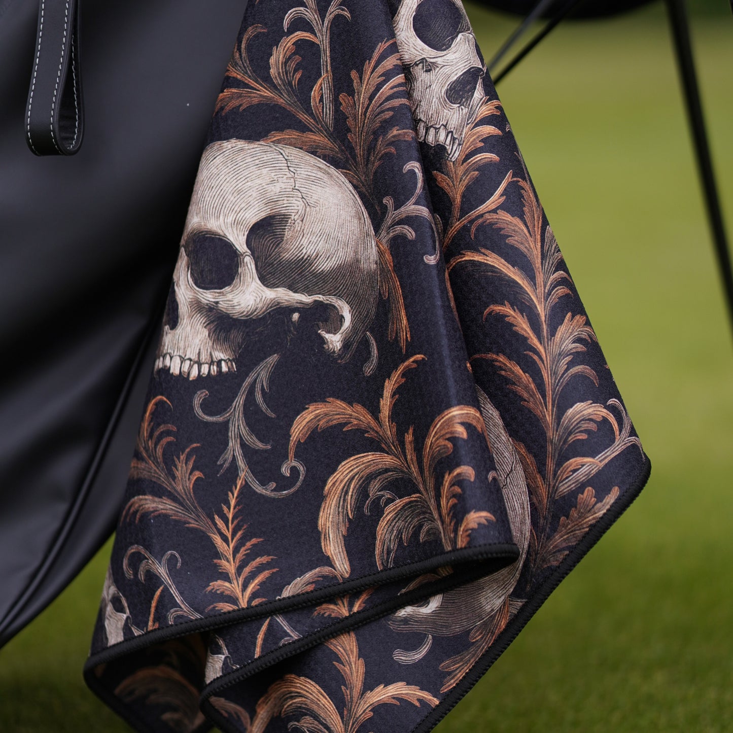 Black golf towel with skull and leaf pattern draped over a black bag on a green background