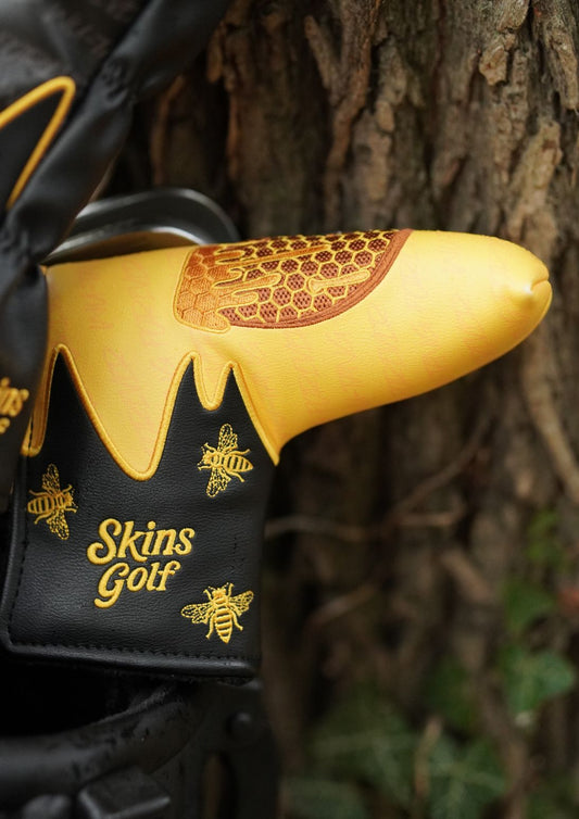 A close-up of a black and yellow golf club headcover with honeycomb and bee designs, featuring the 'Skins Golf' logo, set against a natural background of tree bark.