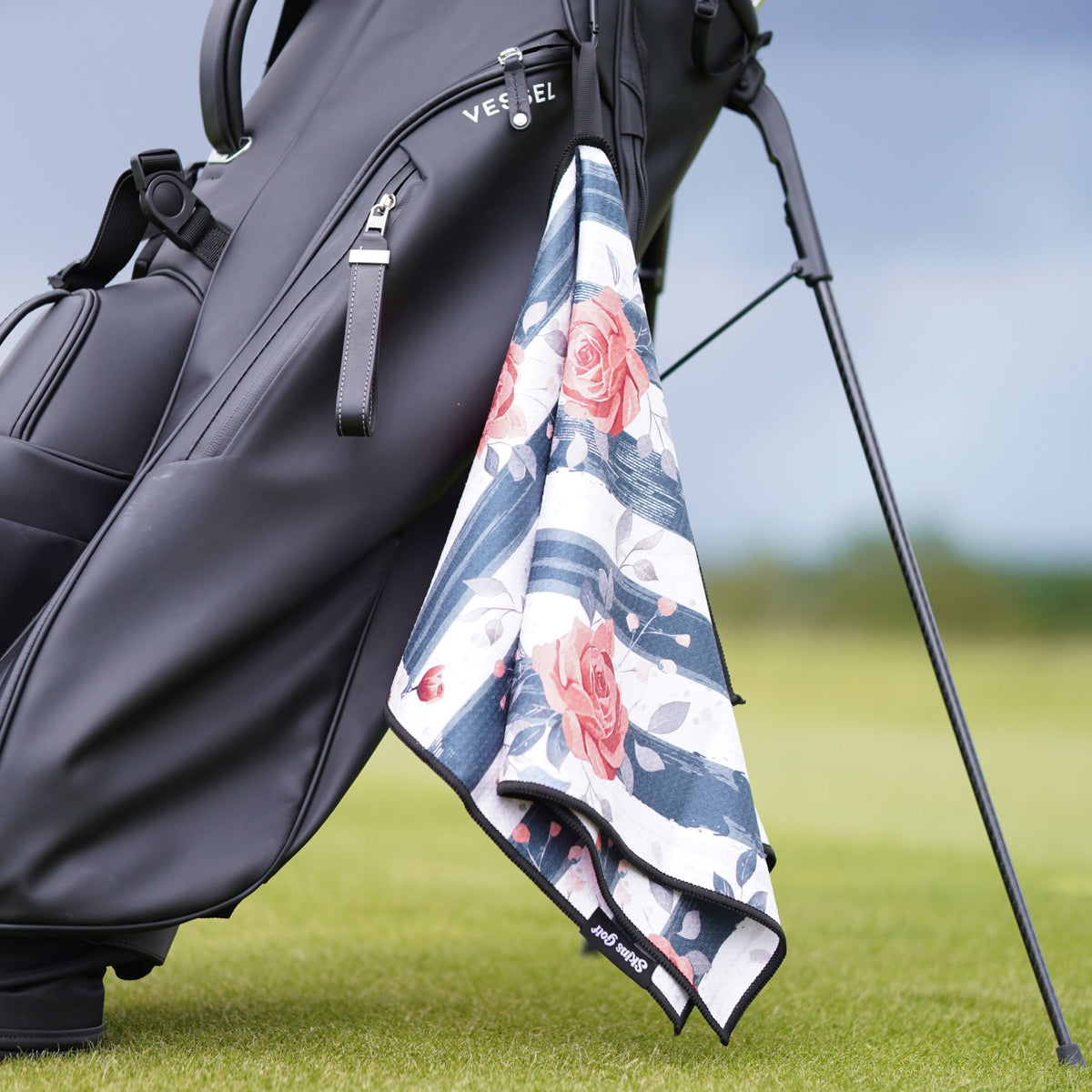 A black golf bag on a golf course with a floral golf towel hanging from the side. The towel features a white base with blue stripes and pink roses.