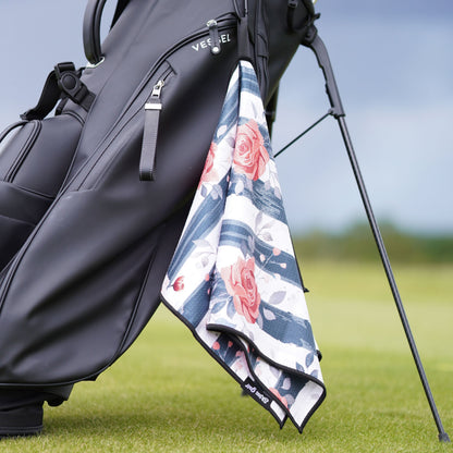 A black golf bag on a golf course with a floral golf towel hanging from the side. The towel features a white base with blue stripes and pink roses.