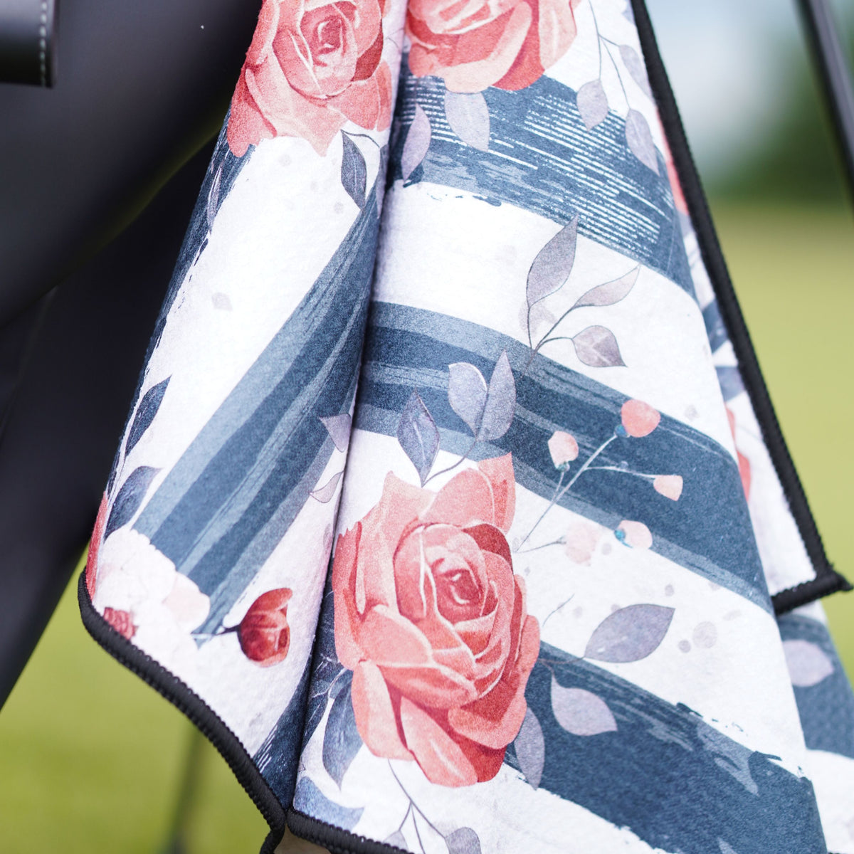 A close-up of a golf towel, featuring a floral print with pink roses, gray leaves, and navy blue brushstroke stripes on a white background.