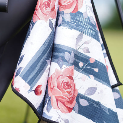 A close-up of a golf towel, featuring a floral print with pink roses, gray leaves, and navy blue brushstroke stripes on a white background.