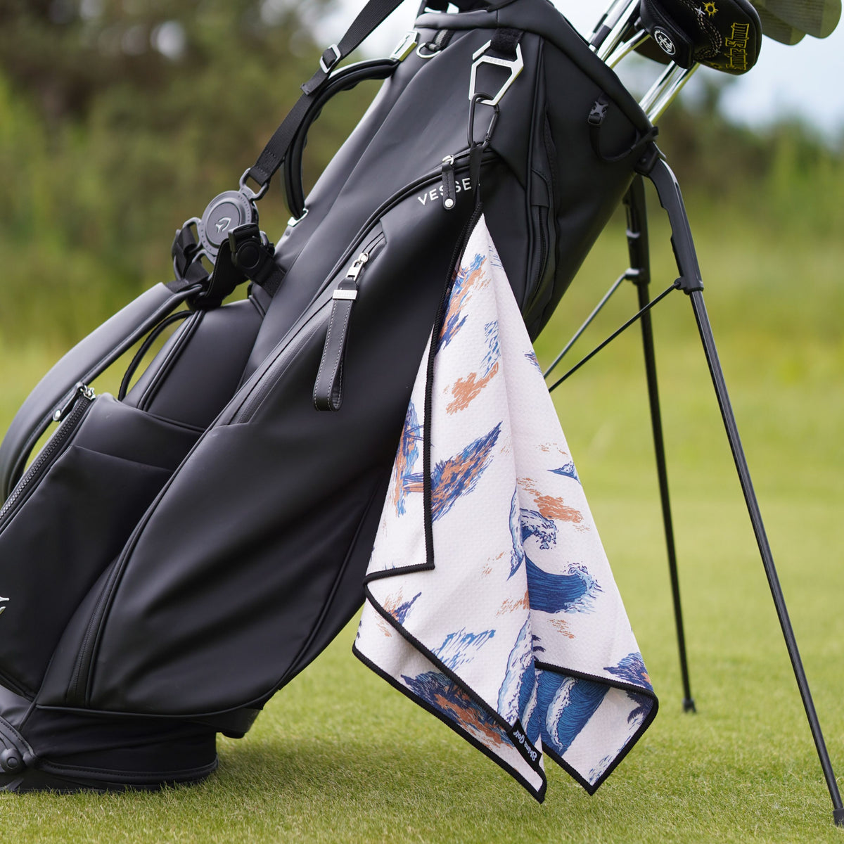 A black golf bag on a grassy golf course with a white hand towel featuring blue and orange brushstroke patterns hanging from the side.