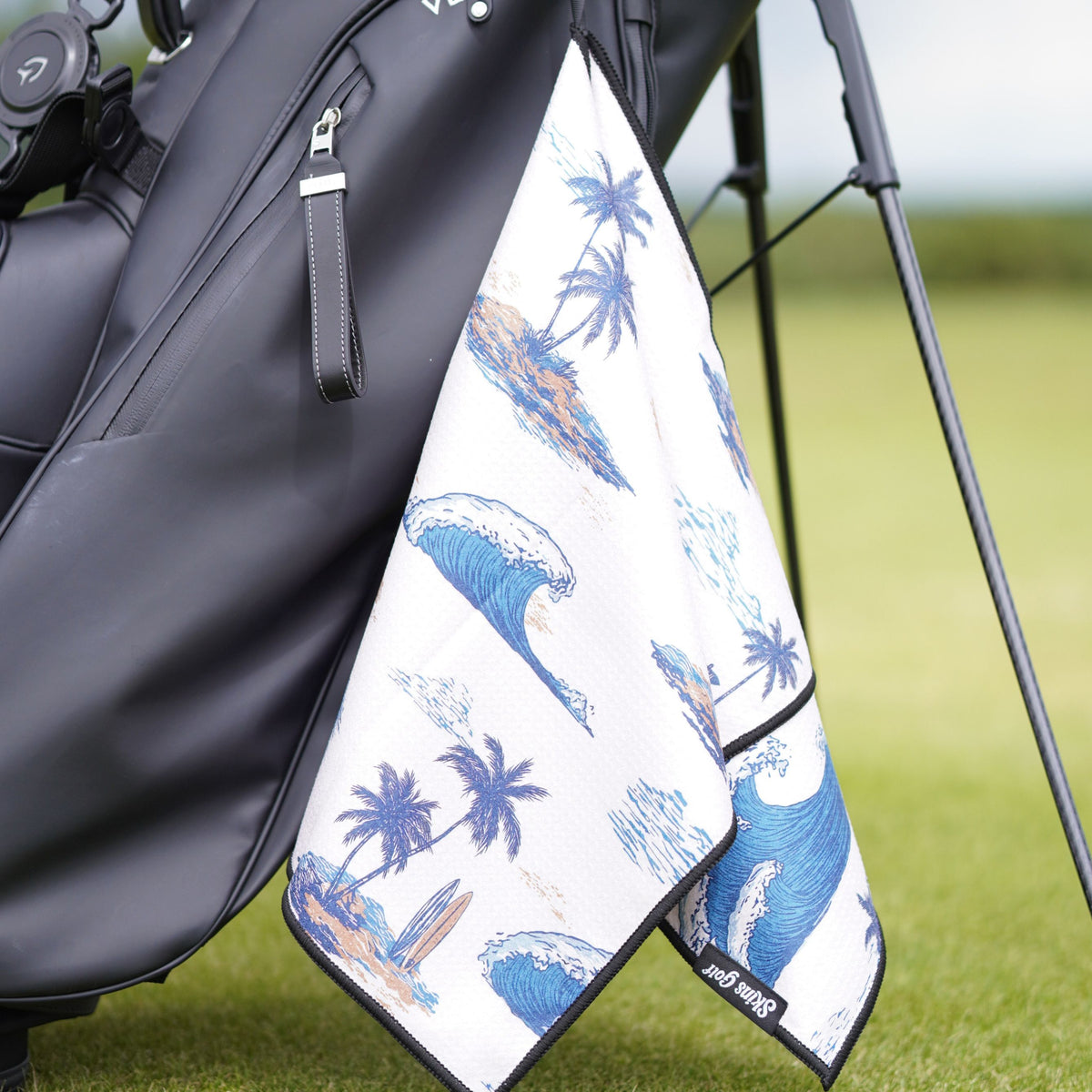 A golf towel with a blue ocean wave and palm tree pattern hanging from a black golf bag on a grassy golf course.