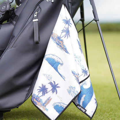 A golf towel with a blue ocean wave and palm tree pattern hanging from a black golf bag on a grassy golf course.