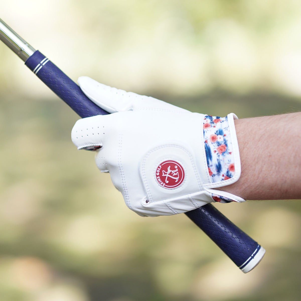 A close-up of a hand holding a golf club, wearing a white golf glove with a red circular logo and a blue, red, and white patterned wrist strap, against a blurred outdoor background.