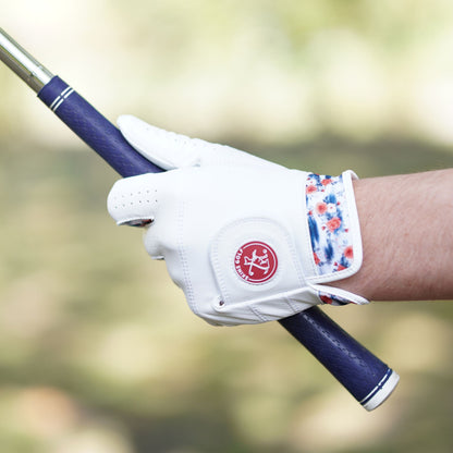 A close-up of a hand holding a golf club, wearing a white golf glove with a red circular logo and a blue, red, and white patterned wrist strap, against a blurred outdoor background.