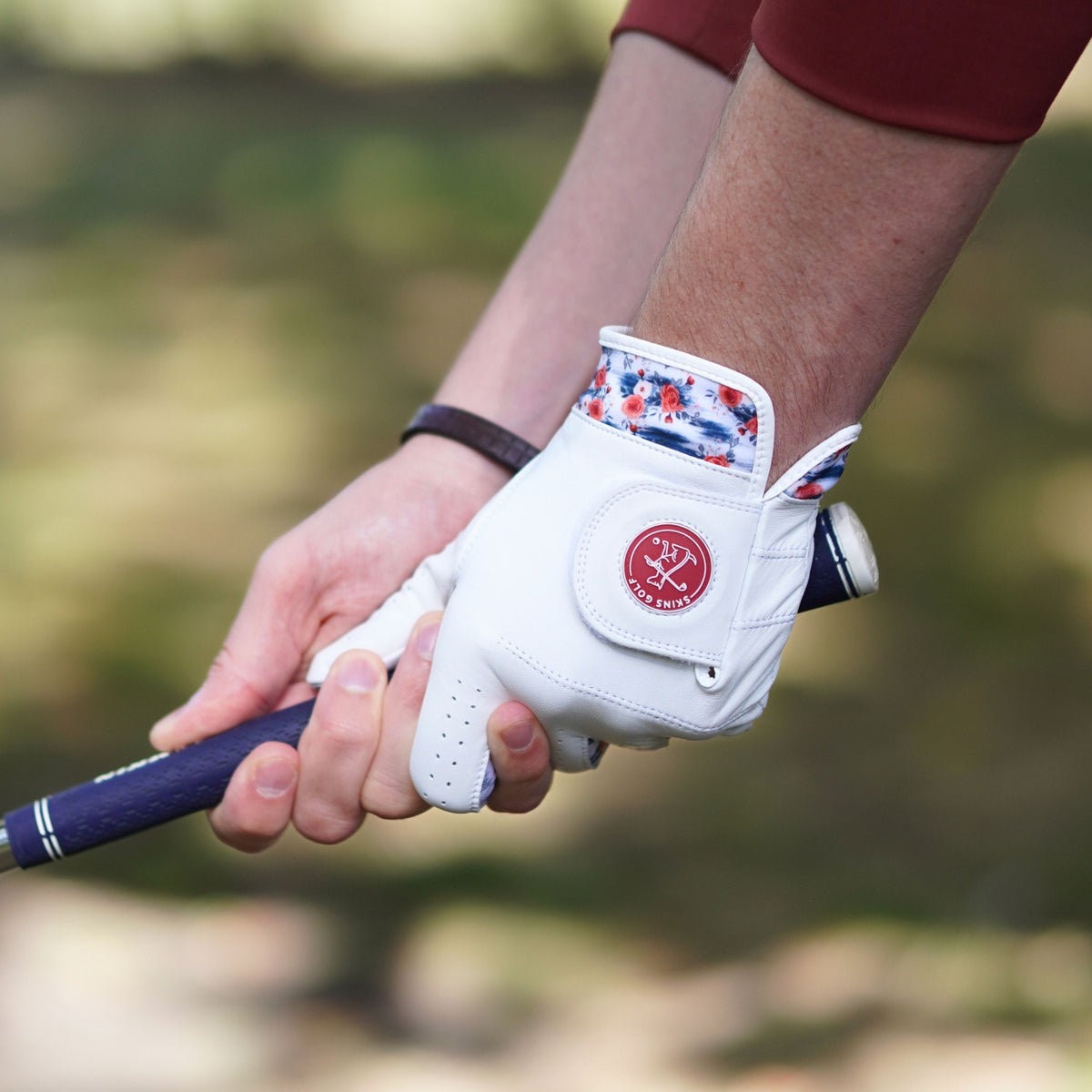 A close-up of a person's hands on a golf club, with the right hand wearing a white golf glove featuring a red circular logo and a patriotic red, white, and blue patterned cuff, against a blurred outdoor background.