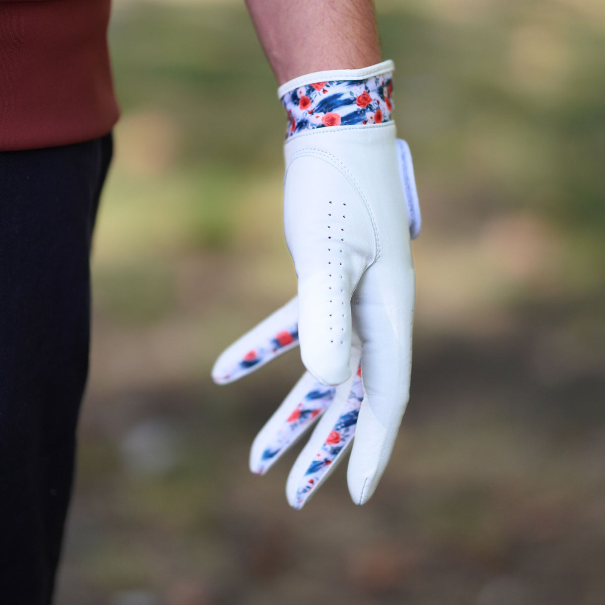 A white golf glove with a red, white, and blue patterned cuff and finger tips, worn on a hand in an outdoor setting.