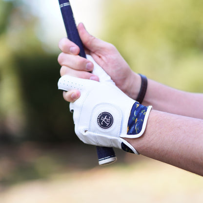 A close-up of a person's hands holding a golf club outdoors, wearing a white golf glove with a navy and gold patterned cuff. The glove features a circular logo patch in the center.