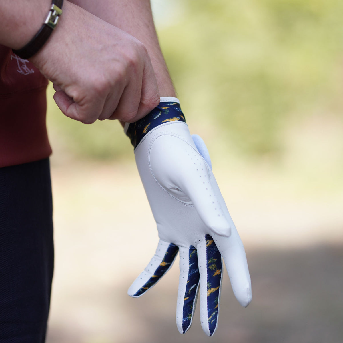 A close-up of a person wearing a white golf glove with navy and gold patterned finger panels and wrist strap, adjusting the cuff. The background is a softly blurred outdoor setting.