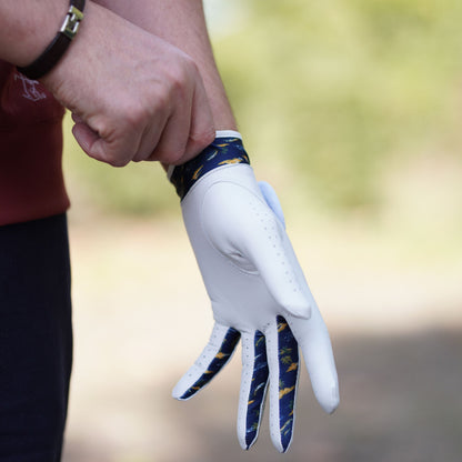 A close-up of a person wearing a white golf glove with navy and gold patterned finger panels and wrist strap, adjusting the cuff. The background is a softly blurred outdoor setting.