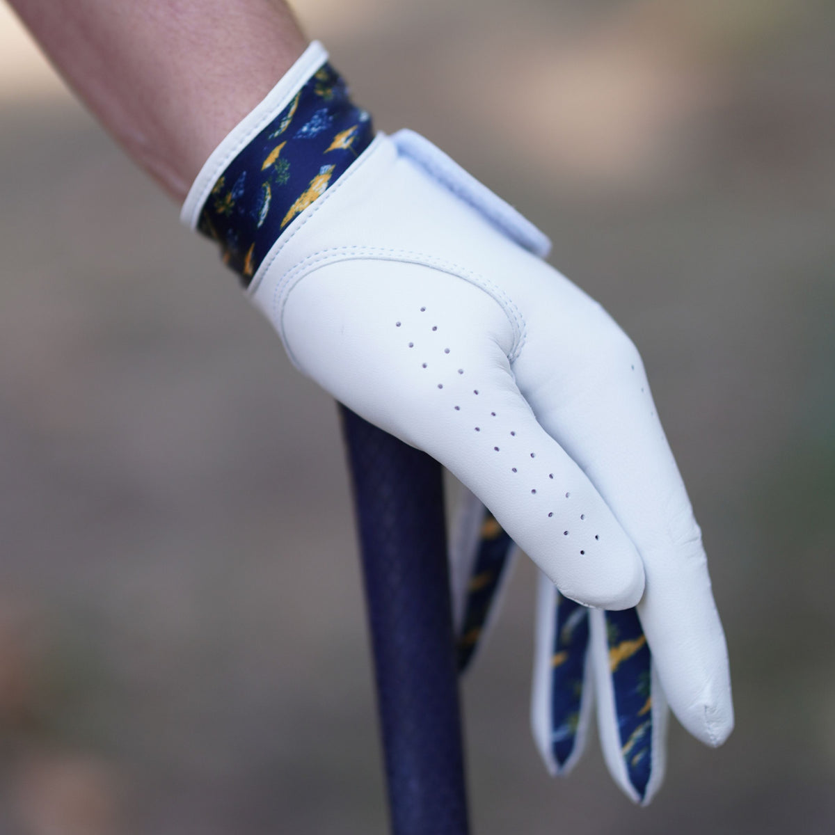A white leather golf glove with a navy and yellow patterned cuff, worn on a hand holding a golf club.