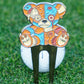 Colorful voodoo bear-shaped golf divot tool in front of a golf ball with grass background