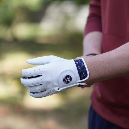 A white golf glove with blue patterned trim and a circular logo patch, worn on a person's hand in an outdoor setting with a blurred green background.
