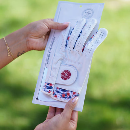 A white golf glove with red and blue floral patterned cuff lining, shown inside its clear retail packaging being held by two hands against a blurred green outdoor background.