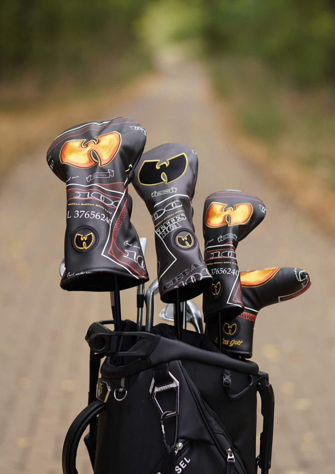 A black golf bag on a blurred outdoor path with three golf club headcovers standing out, featuring a gold and black Wu-Tang Clan logo design.