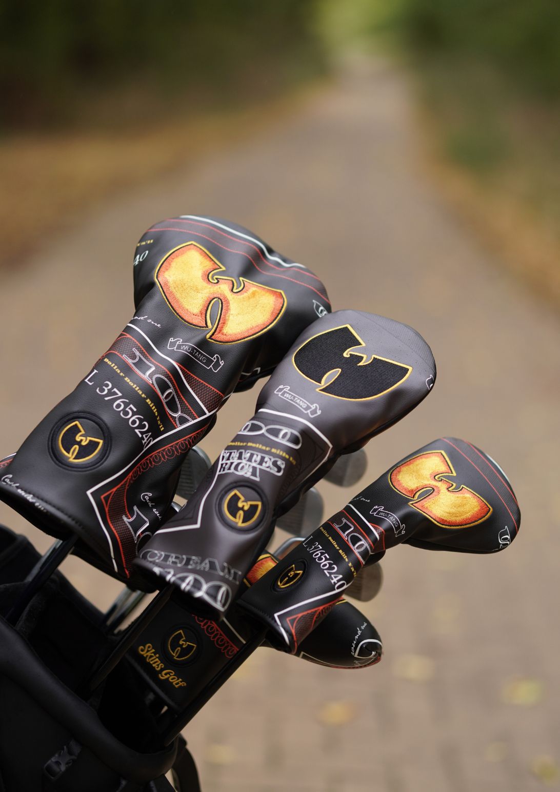Three golf club headcovers with a black and gold Wu-Tang Clan design, resting in a black golf bag on a blurred path.