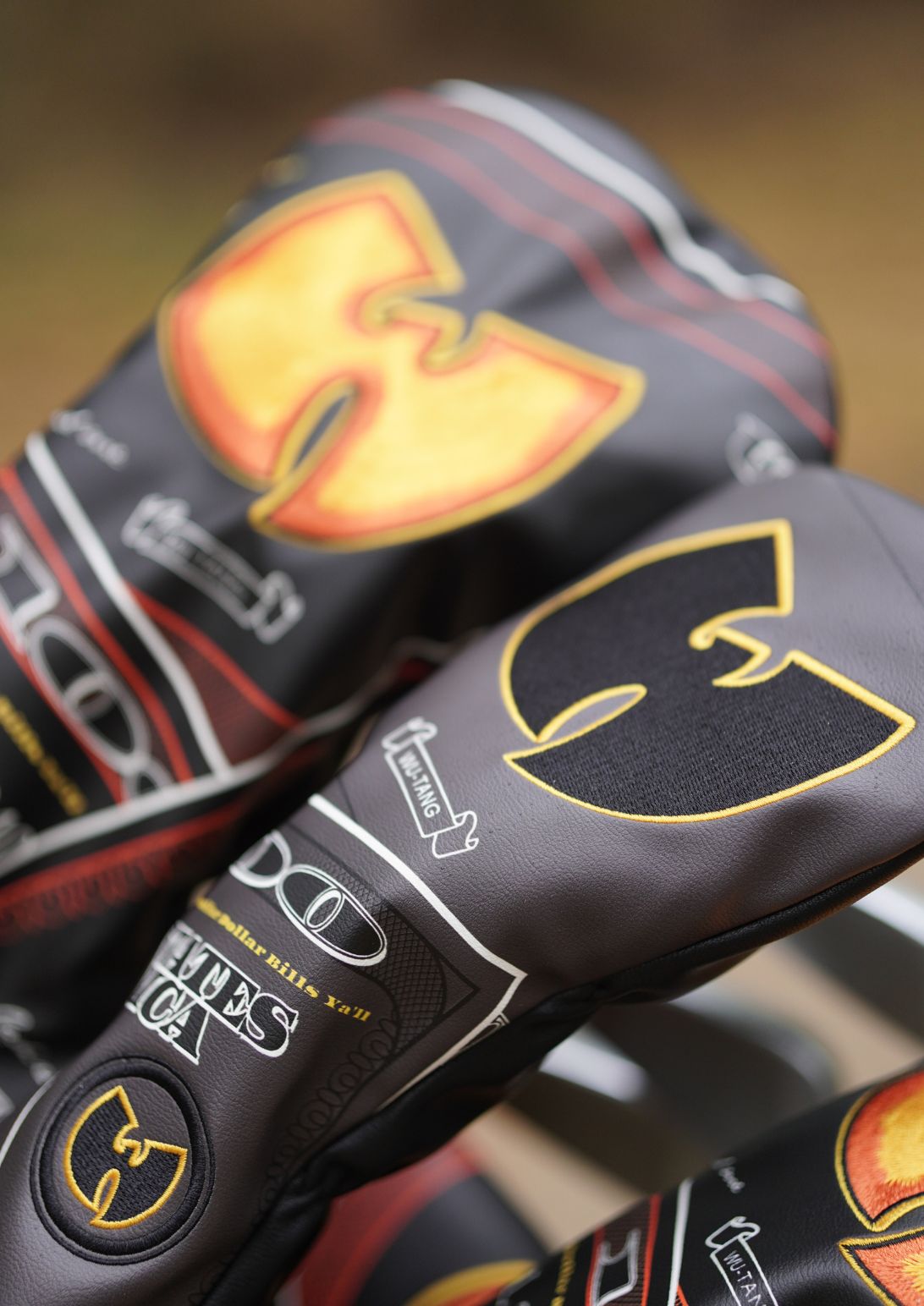 Close-up of a golf club headcover with a black background, a large gold and orange Wu-Tang Clan logo, and additional printed graphics and text.