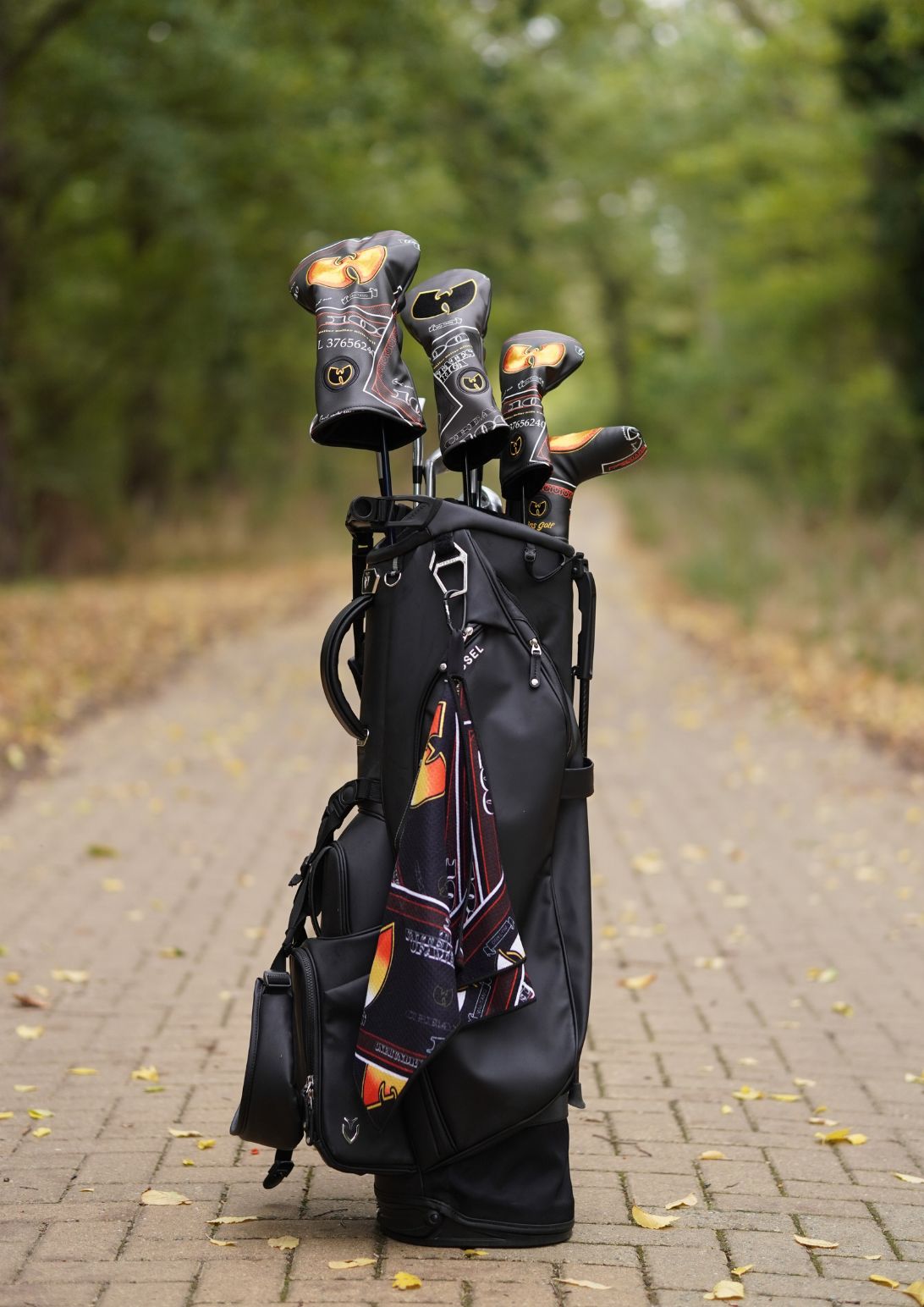 A black golf bag standing on a paved path outdoors, holding several golf clubs with decorative black, orange, and gold headcovers. Trees and fallen leaves are visible in the blurred background.