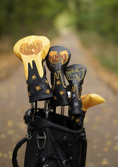 A golf bag on a blurred outdoor path with four custom black and gold golf club headcovers sticking out. The headcovers feature a honeycomb pattern, dripping wax design, and bee motifs.