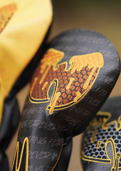 Close-up of a golf club headcover in black with a large gold and orange honeycomb-style graphic and gold embroidered lettering, alongside partial views of yellow and black headcovers in the background.