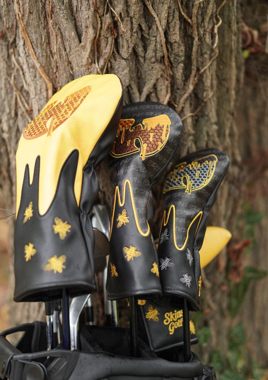 A set of golf club headcovers in black and yellow, resting in a golf bag against a tree trunk outdoors.