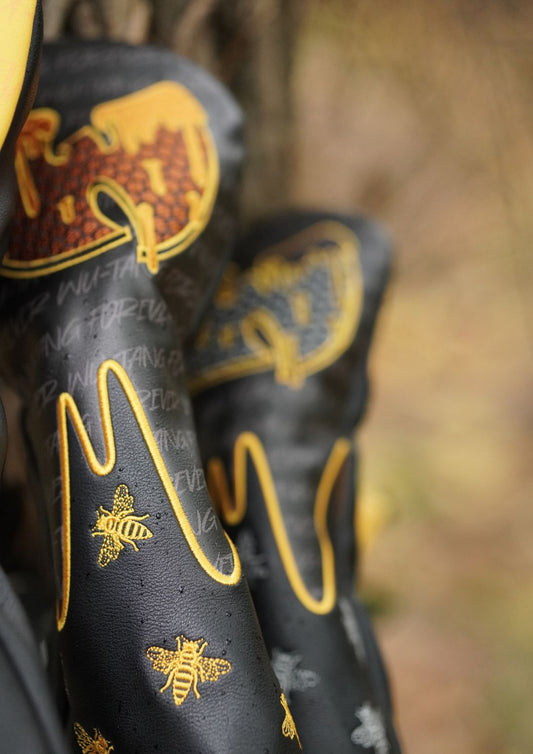 Close-up of black golf club covers with gold bee and honeycomb designs, photographed outdoors against a blurred natural background.
