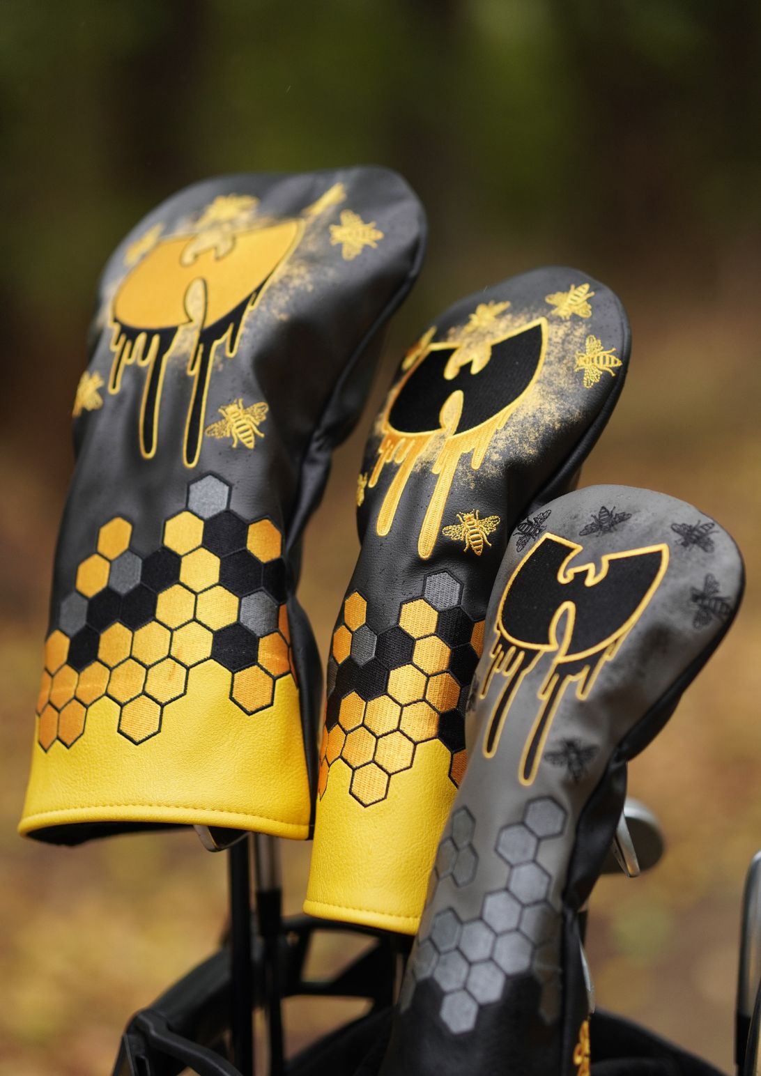 Three golf club headcovers with a honeycomb and bee design in black and yellow, shown standing in a bag outdoors with a blurred forest background.