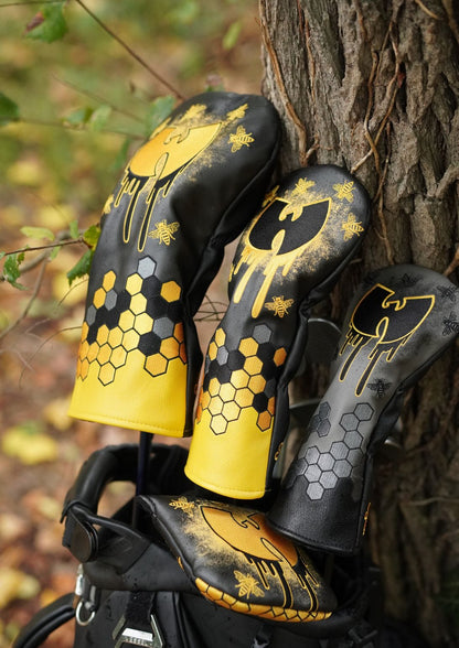 Three black and yellow golf club headcover sleeves with gold honeycomb and graffiti-style graphics, leaning against a tree outdoors.