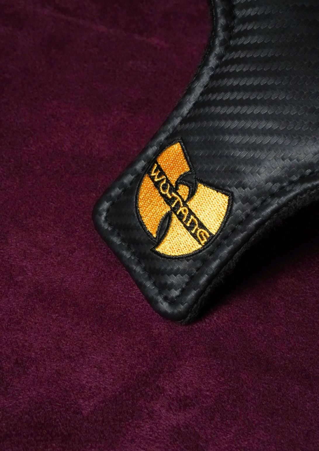 A close-up of a black carbon-fiber-textured leather mallet headcover with an embroidered gold and black Wu-Tang Clan logo patch, set against a deep purple textured background.