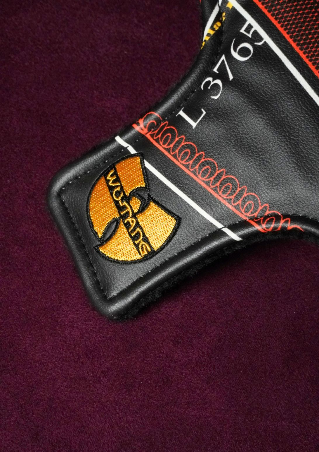 Close-up of a black leather golf headcover featuring an embroidered yellow and black Wu-Tang Clan patch, white numbering, and orange-red stitched details, set against a dark purple textured background.