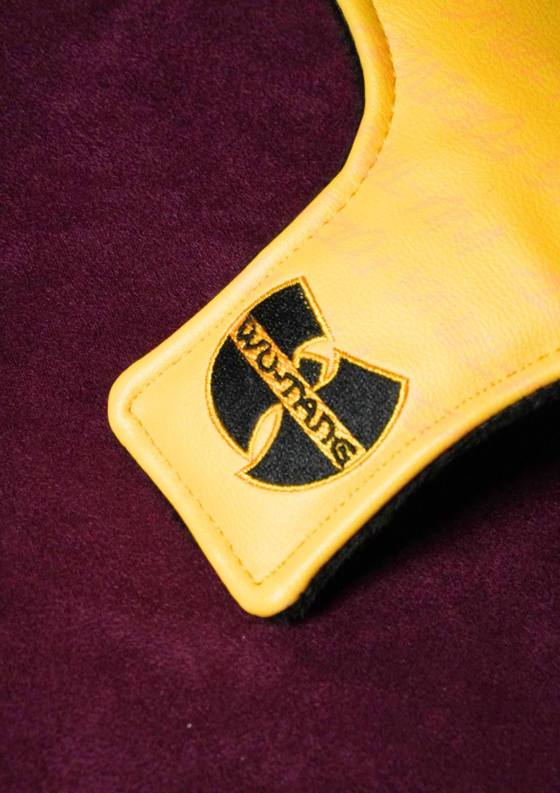A close-up of a yellow leather mallet headcover with a black embroidered Wu-Tang Clan logo patch on a deep burgundy background.