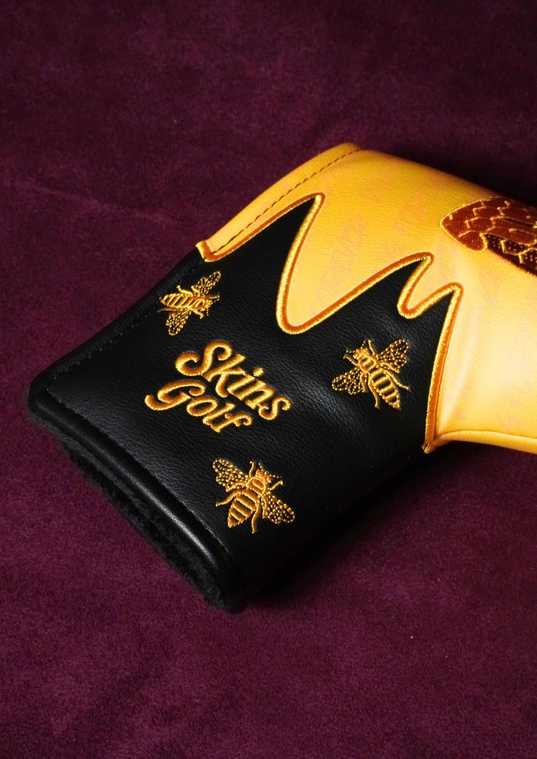 A close-up of a golf blade headcover on a deep purple velvet background. The headcover features a black leather lower section with gold embroidered bees and the text 'Skins Golf', a gold honeycomb pattern, and a gold honeycomb-shaped upper section.