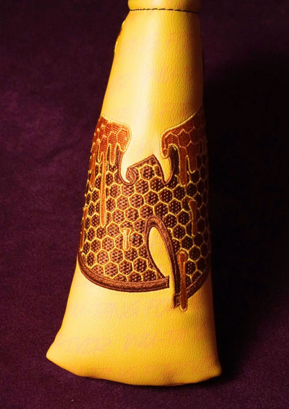 A close-up of a yellow leather blade headcover featuring a brown embroidered honeycomb and abstract design on the side, set against a dark purple background.