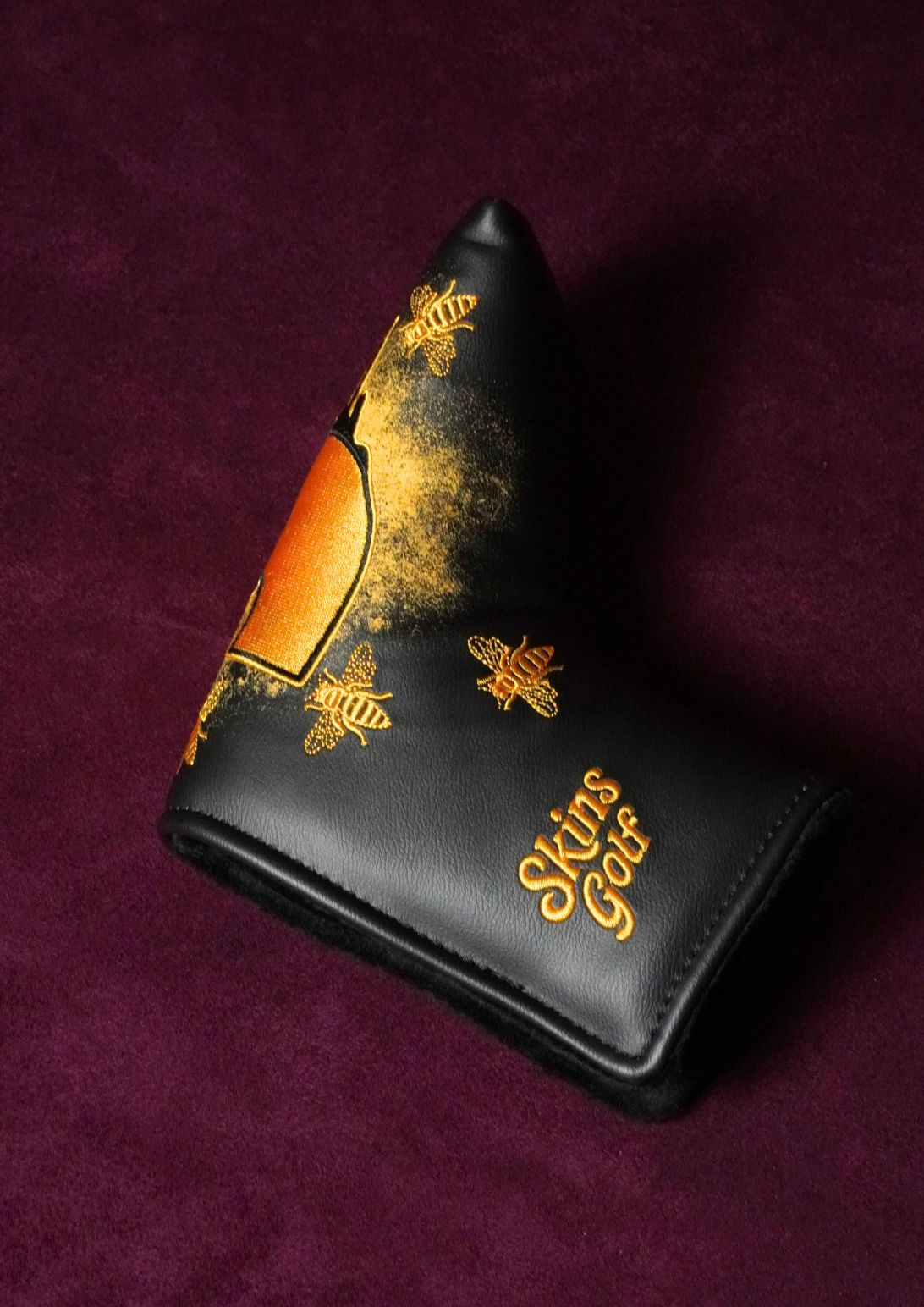 A black leather golf putter headcover with gold bee designs and the text 'Skins Golf' on a dark purple textured background.