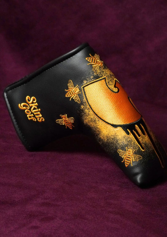 A black leather putter headcover with gold spray-painted bee designs and a gold shield logo, displayed on a textured dark purple background.