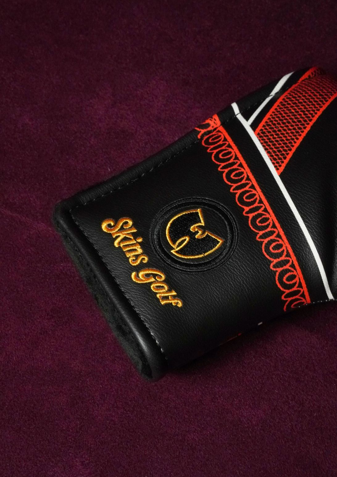 A close-up of a black leather golf club headcover with gold and orange embroidered details, including the text 'Skins Golf' and a circular logo, resting on a textured dark purple background.