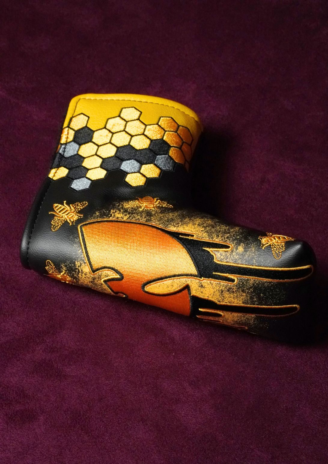 A custom golf putter headcover featuring a black and gold honeycomb pattern on the top, a large gold graphic on the front, and gold bee designs, displayed on a dark purple textured background.