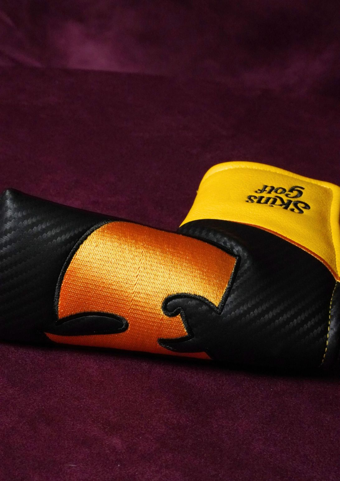 A close-up of a blade headcover featuring a black carbon-fiber pattern, a central orange and black wu-tang clan logo, and a bright yellow cuff with the brand name 'Skins Golf' printed in black.