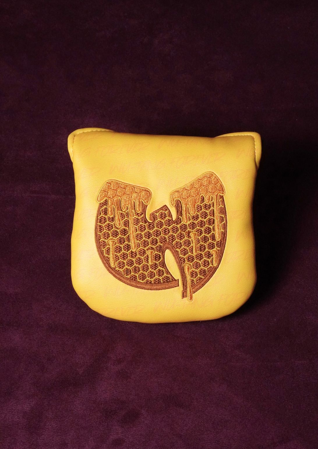 A yellow leather mallet headcover featuring a brown engraved Wu-Tang Clan logo, set against a dark purple textured background.