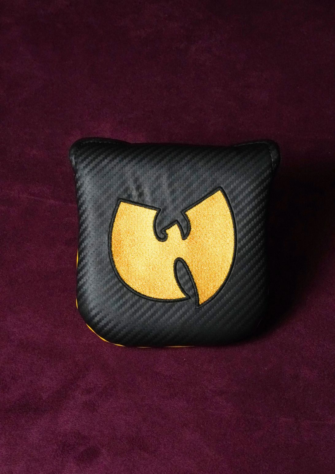A square black carbon-fiber-textured golf mallet headcover featuring a large gold Wu-Tang Clan logo on the front, set against a dark purple textured background.