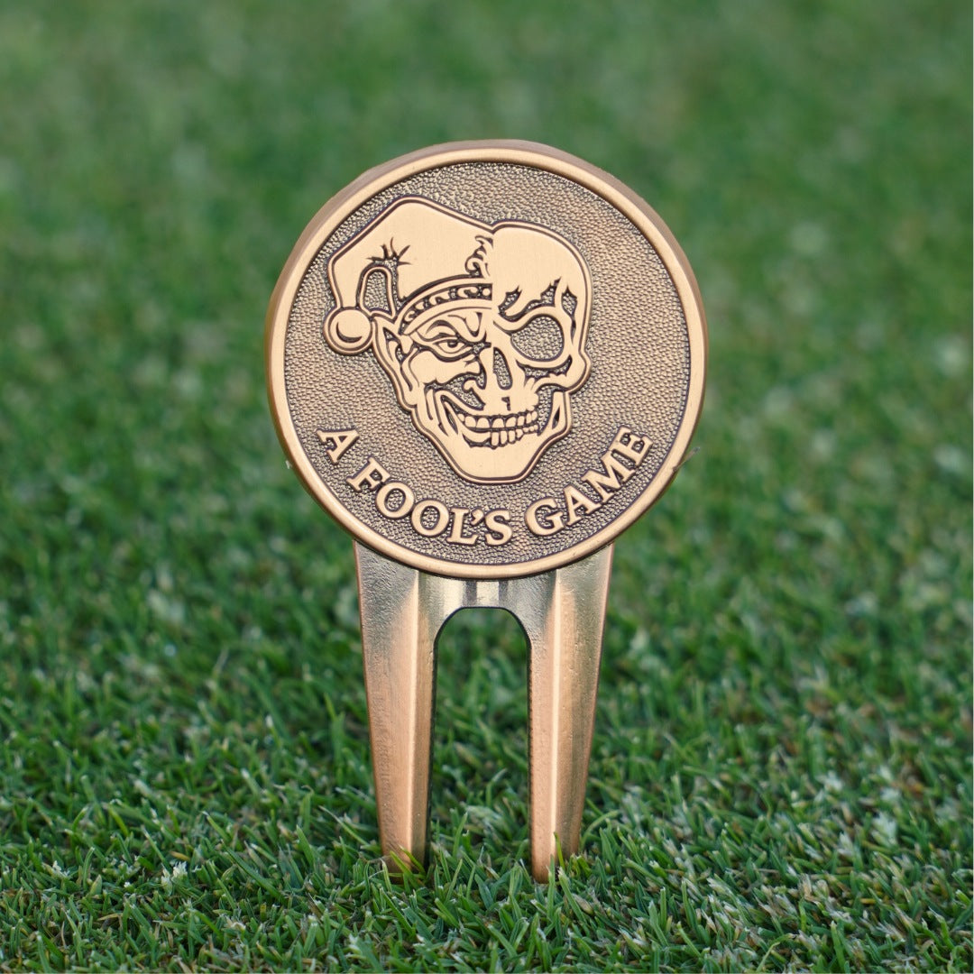 Bronze golf divot tool with skull design and 'A Fool's Game' text on grass