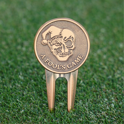 Bronze golf divot tool with skull design and 'A Fool's Game' text on grass